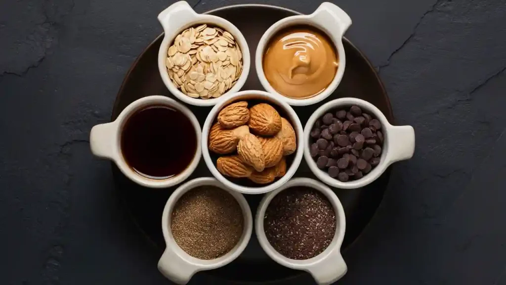 Flat lay of energy bites ingredients including oats, nut butter, honey, chia seeds, chocolate chips, and vanilla extract arranged on a kitchen counter.